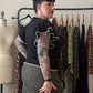 A woman with dark hair in a bun and colorful arm tattoos stands in profile next to a clothing rack, wearing a black short-sleeve top with white ruffle trim and a green leopard-print skirt. A cream dress form is visible on the left against a plain beige wall.