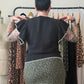 A person viewed from behind wearing a black ruffled-sleeve top with white trim and a leopard-print skirt, standing in front of a clothing rack in a studio setting.