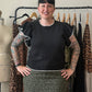 A smiling woman with short black hair and visible arm tattoos stands in front of a clothing rack, wearing a black short-sleeve top with white ruffle trim and a green leopard-print skirt. A mannequin and other garments are visible in the background.