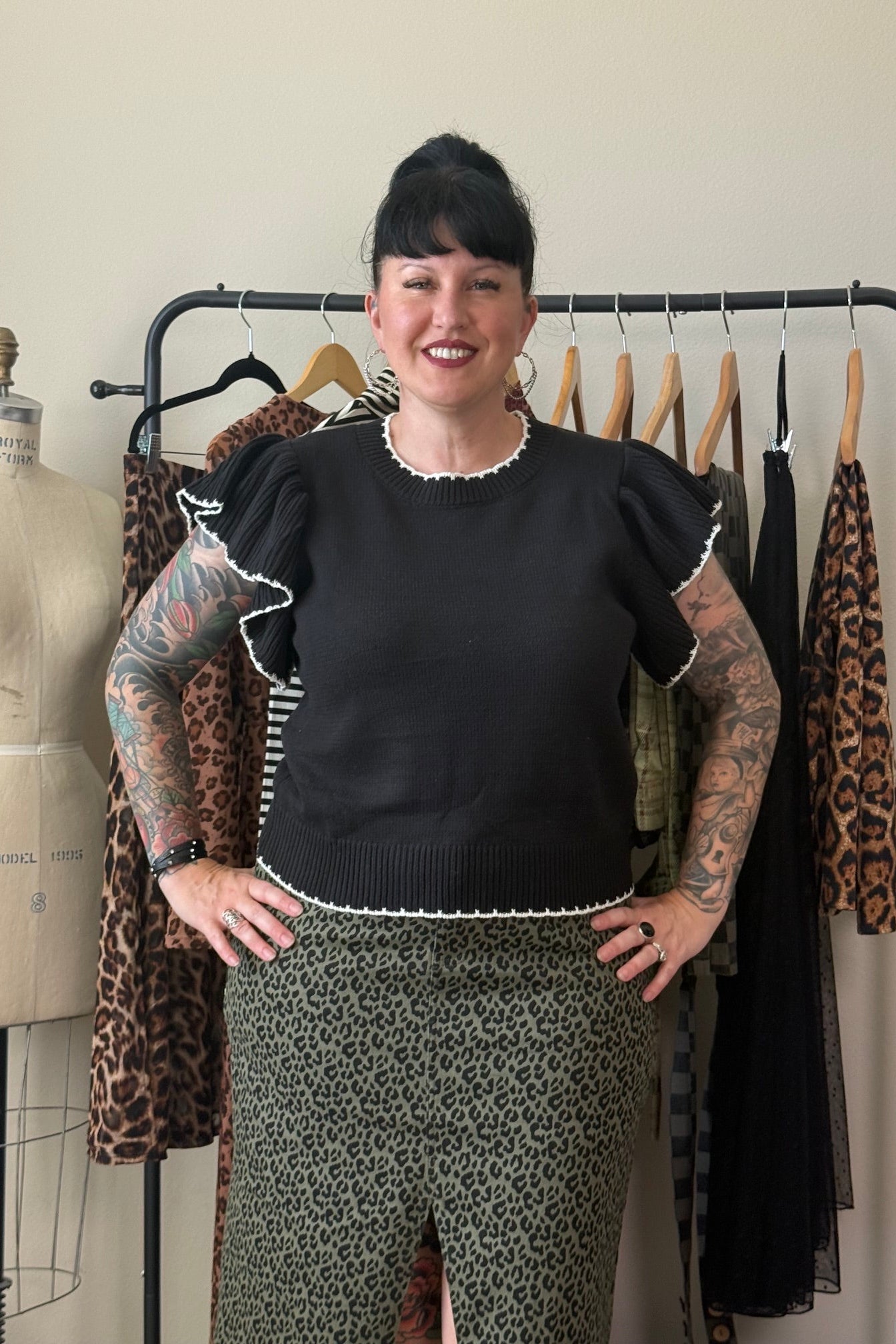 A smiling woman with short black hair and visible arm tattoos stands in front of a clothing rack, wearing a black short-sleeve top with white ruffle trim and a green leopard-print skirt. A mannequin and other garments are visible in the background.