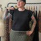 A woman with tattoos models a black short-sleeve knit top with white lace trim and a green leopard-print skirt, standing in front of a clothing rack.