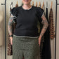 A woman with black hair and tattoos stands in front of a clothing rack, wearing a black short-sleeve top with ruffled trim and olive green leopard-print pants. A dress form is visible on the left, and several garments hang on a black rack behind her.