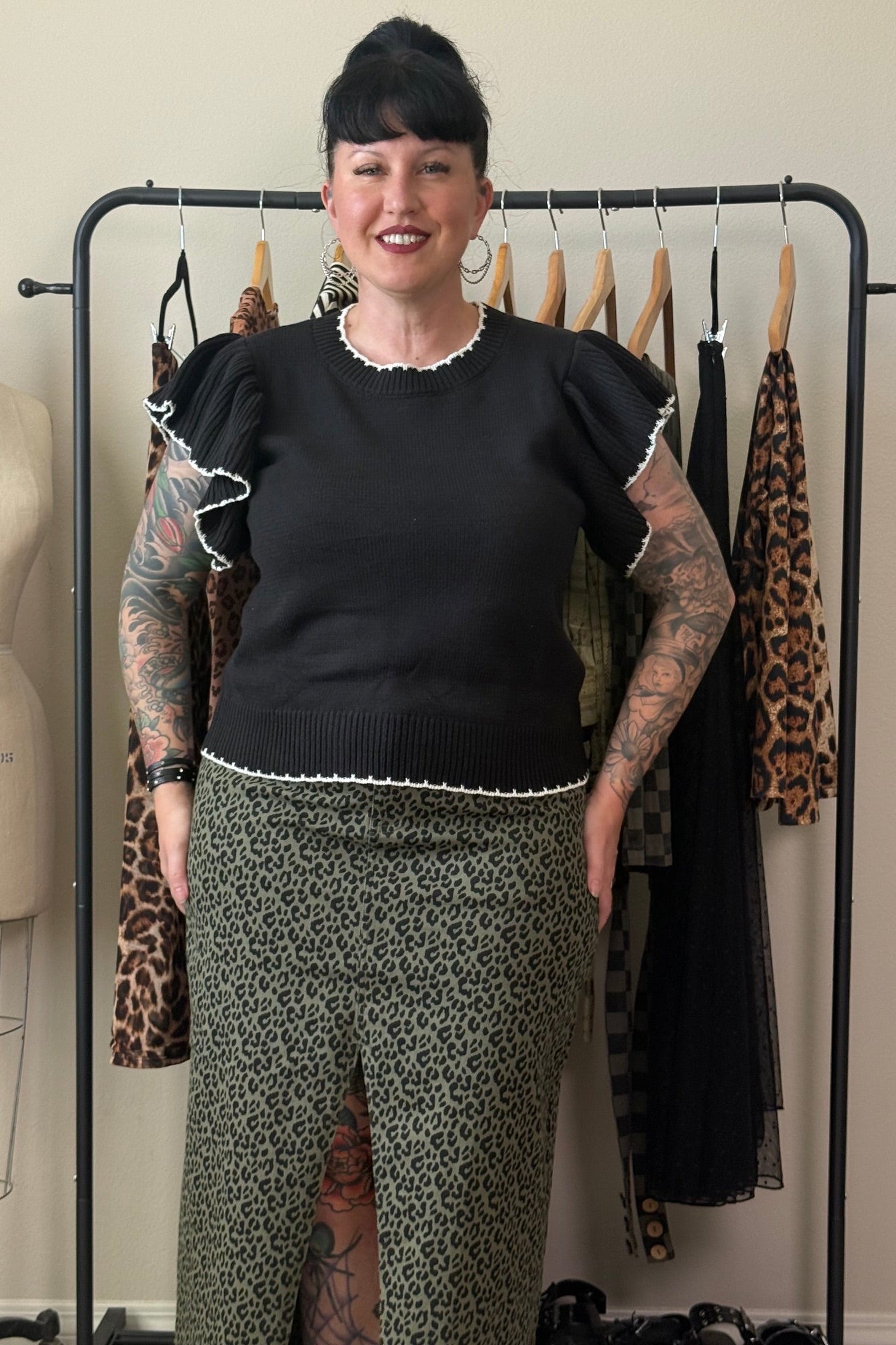 A woman with black hair and tattoos stands in front of a clothing rack, wearing a black short-sleeve top with ruffled trim and olive green leopard-print pants. A dress form is visible on the left, and several garments hang on a black rack behind her.
