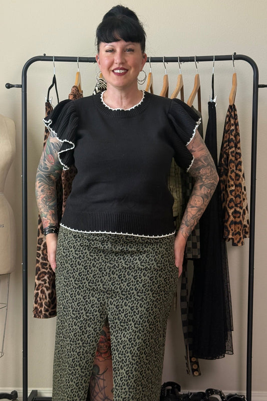 A woman with black hair and tattoos stands in front of a clothing rack, wearing a black short-sleeve top with ruffled trim and olive green leopard-print pants. A dress form is visible on the left, and several garments hang on a black rack behind her.