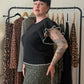 A woman with short black hair and arm tattoos stands in front of a clothing rack, wearing a black ruffled short-sleeve top and a green leopard-print skirt. A mannequin and other garments are visible in the background.
