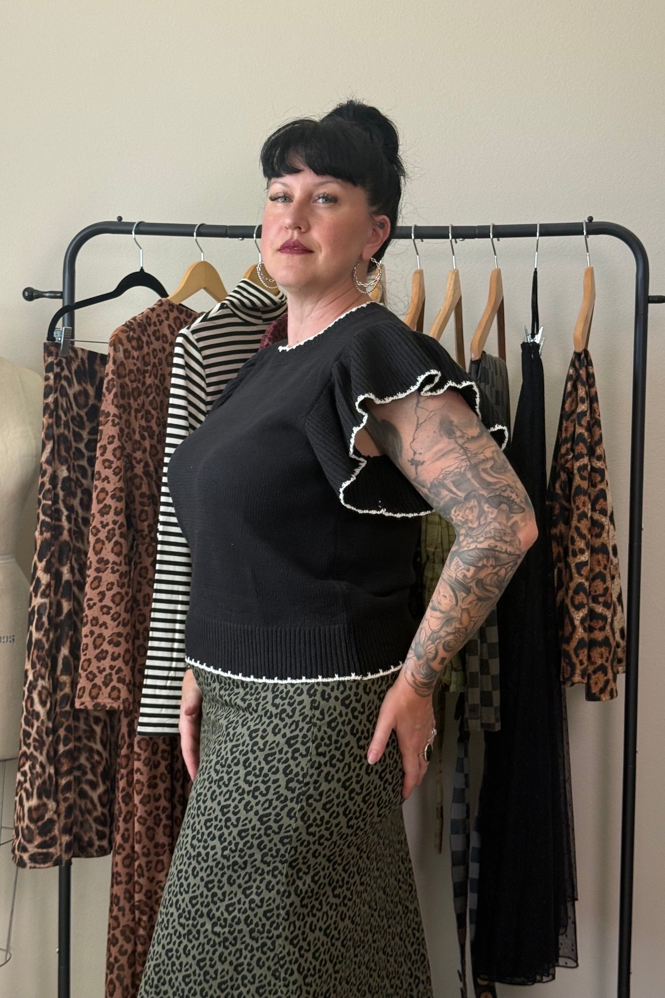 A woman with short black hair and arm tattoos stands in front of a clothing rack, wearing a black ruffled short-sleeve top and a green leopard-print skirt. A mannequin and other garments are visible in the background.