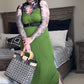 A person wearing a green dress and black boots, holding a patterned bag, with a harness around the neck.