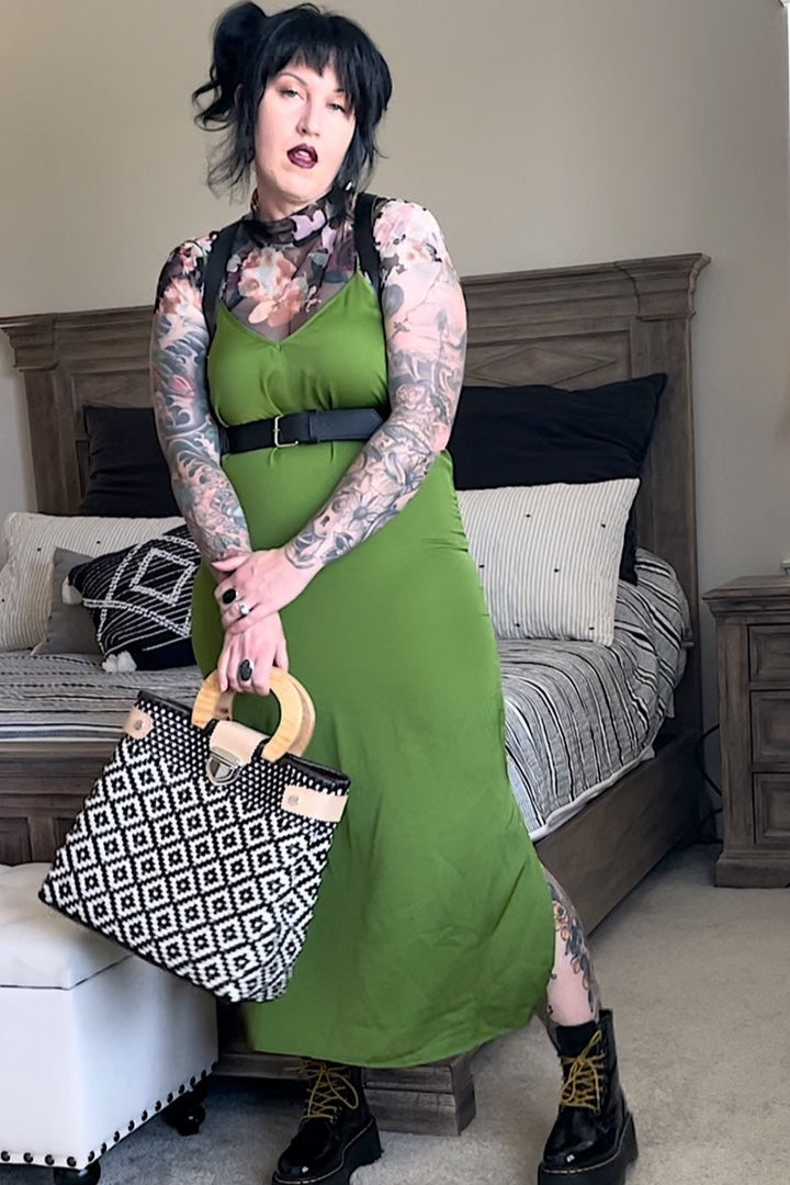 A person wearing a green dress and black boots, holding a patterned bag, with a harness around the neck.