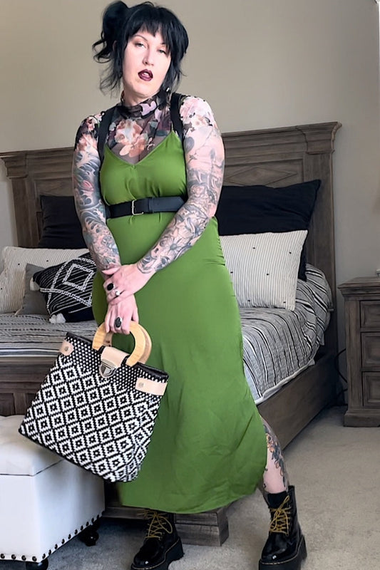 A person wearing a green dress and black boots, holding a patterned bag, with a harness around the neck.