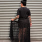 A person stands with their back to the camera in front of a gray metal roll-up door, wearing a black short-sleeve top and a long sheer black mesh skirt. The person is holding out the side of the skirt to show its drape and texture.