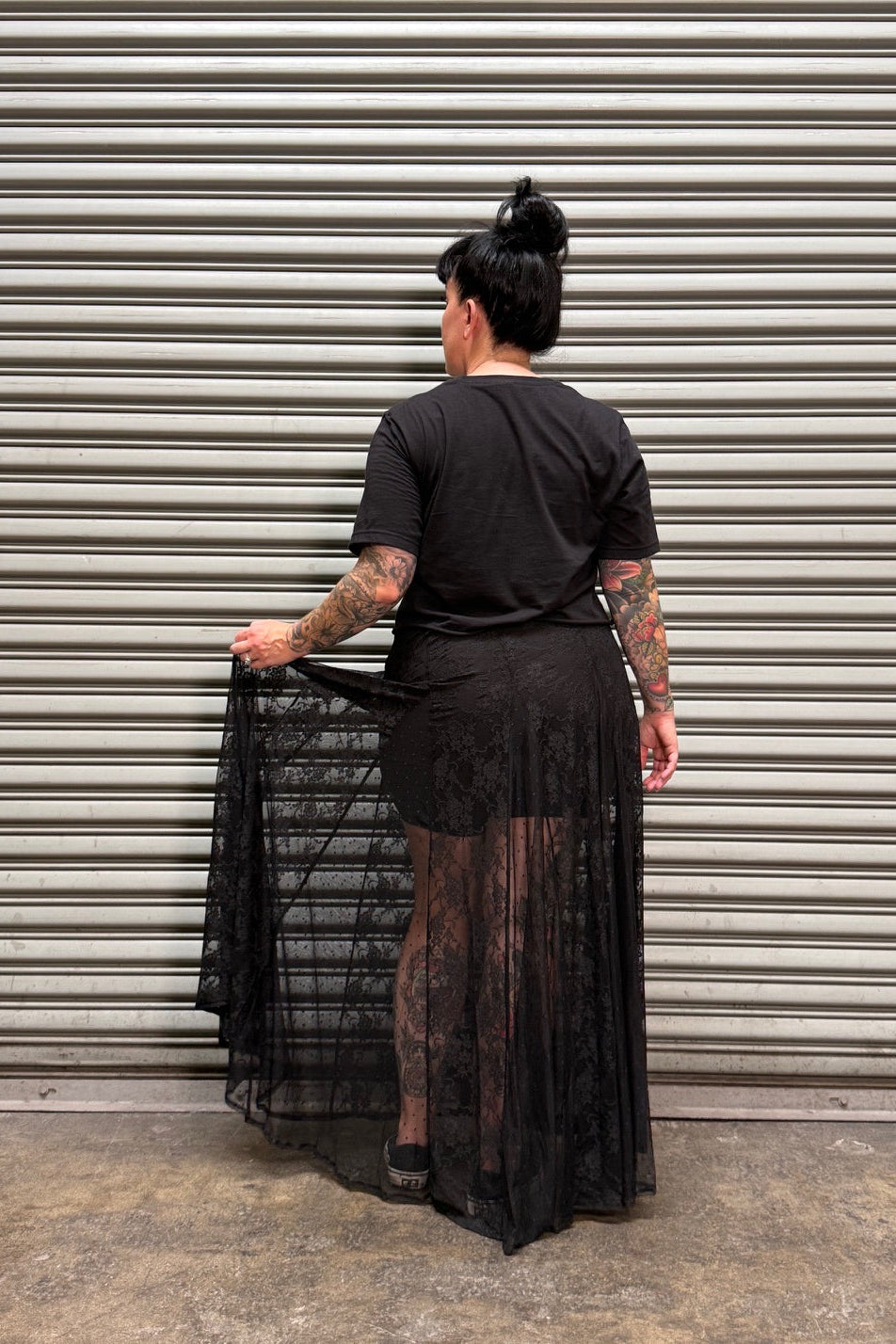 A person stands with their back to the camera in front of a gray metal roll-up door, wearing a black short-sleeve top and a long sheer black mesh skirt. The person is holding out the side of the skirt to show its drape and texture.