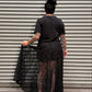 Woman with black hair and tattoos showing her backside standing in front of a metal backdrop.