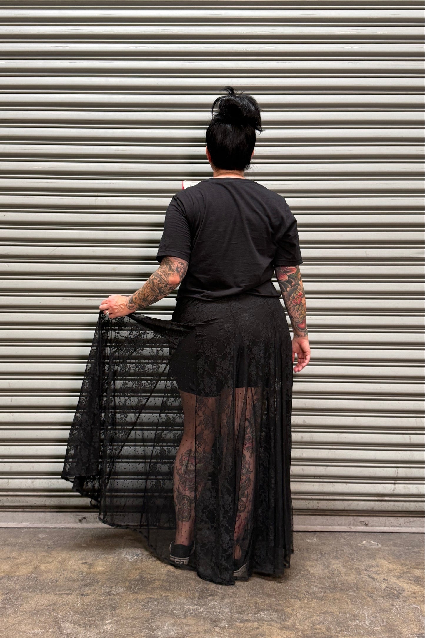 Woman with black hair and tattoos showing her backside standing in front of a metal backdrop.