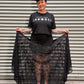 Woman with black hair and tattoos wearing a moon phase crop top and a lace maxi skirt while holding up her skirt in front of a metal back drop.