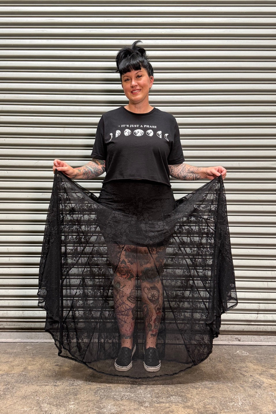 Woman with black hair and tattoos wearing a moon phase crop top and a lace maxi skirt while holding up her skirt in front of a metal back drop.