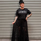 Woman with black hair and tattoos wearing a black crop top and maxi skirt standing in front of a metal back drop