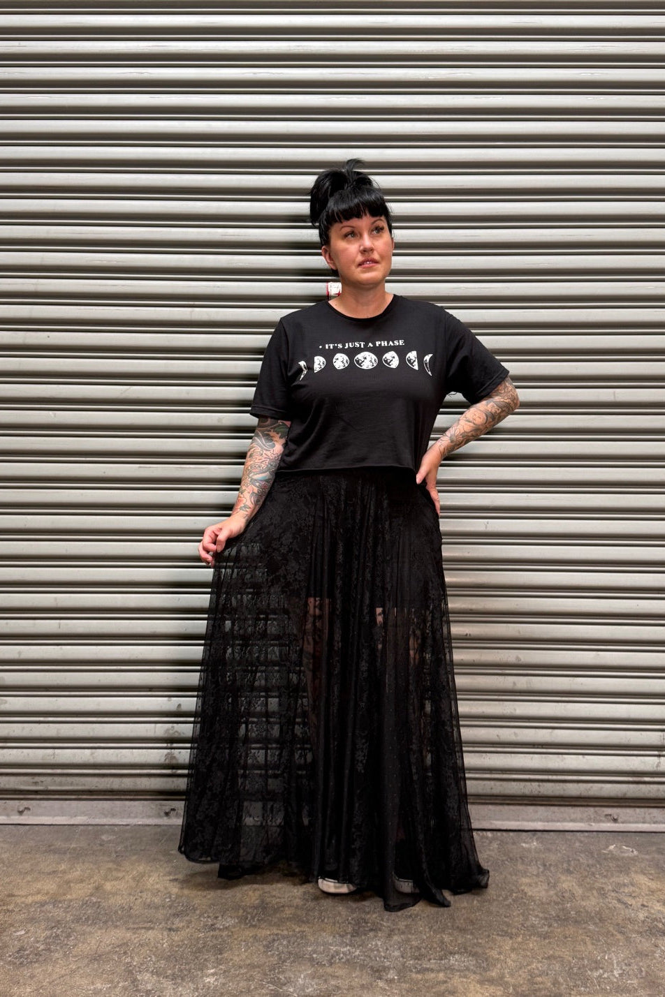 Woman with black hair and tattoos wearing a black crop top and maxi skirt standing in front of a metal back drop