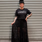 Woman with black hair and tattoos wearing a moon phase crop top and a lace maxi skirt in front of a metal backdrop.