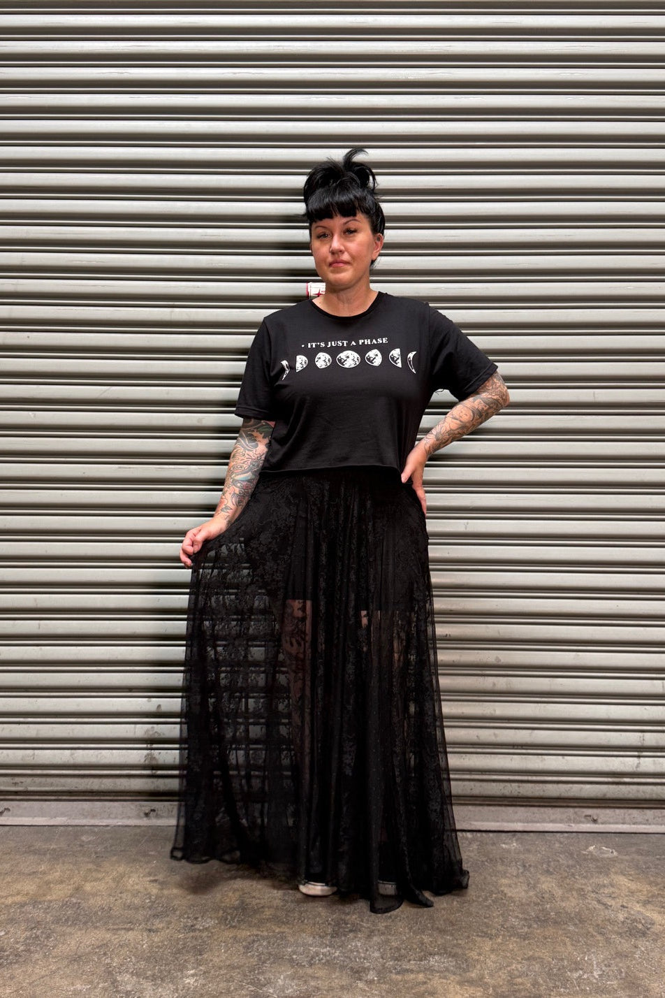 Woman with black hair and tattoos wearing a moon phase crop top and a lace maxi skirt in front of a metal backdrop.