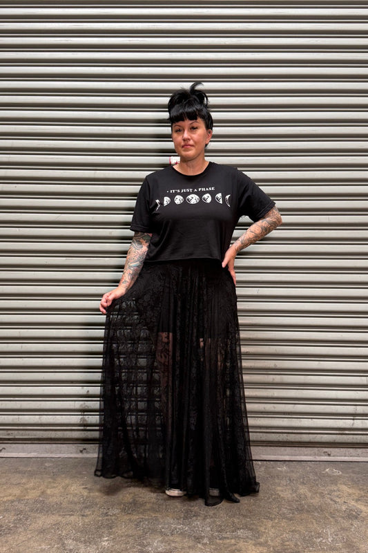 Woman with black hair and tattoos wearing a moon phase crop top and a lace maxi skirt in front of a metal backdrop.