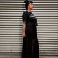 A woman with dark hair in a bun stands in profile against a gray metal roll-up door, wearing a black short-sleeve graphic T-shirt and a long black pleated skirt with sheer lace overlay.