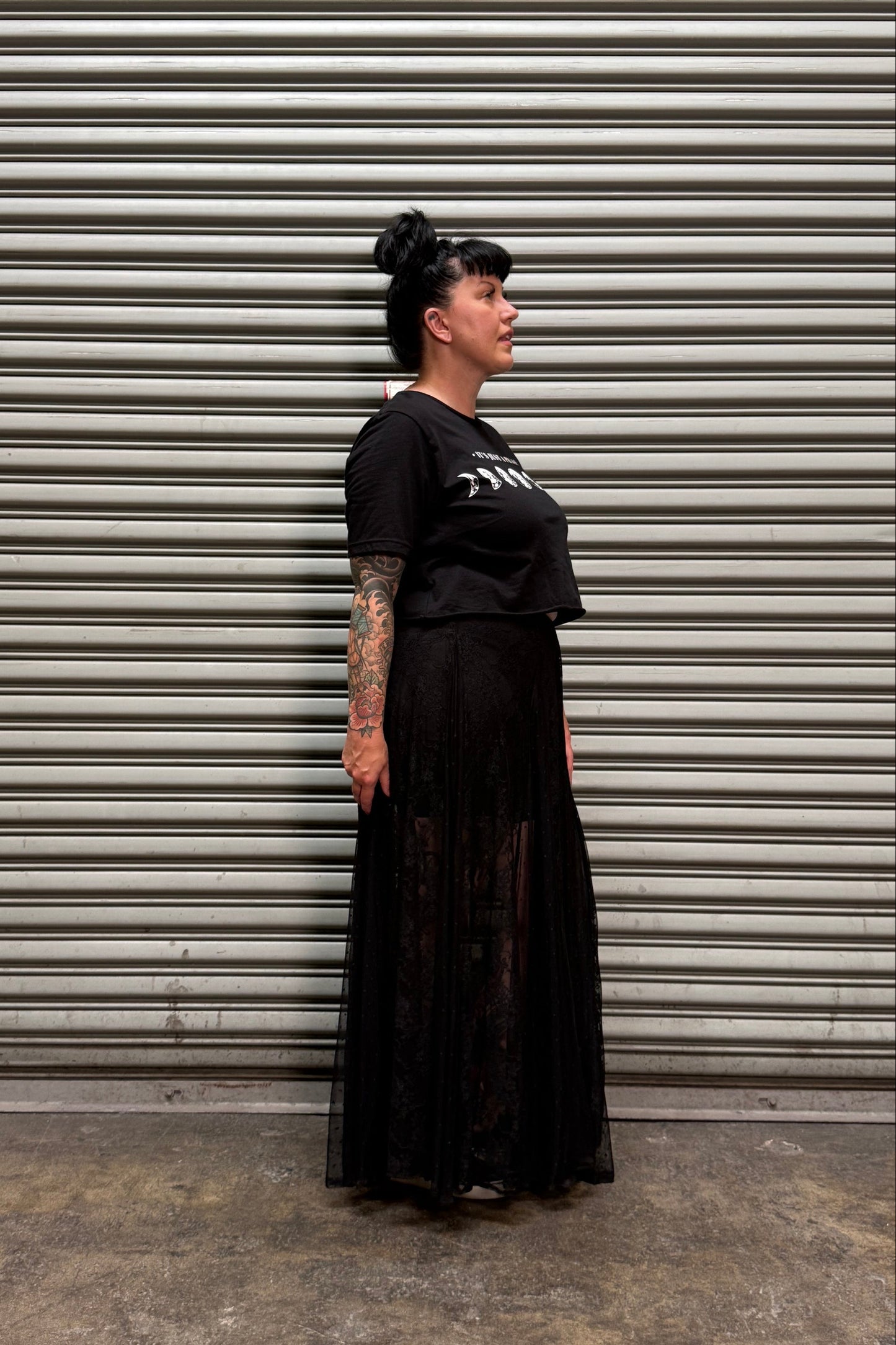 A woman with dark hair in a bun stands in profile against a gray metal roll-up door, wearing a black short-sleeve graphic T-shirt and a long black pleated skirt with sheer lace overlay.