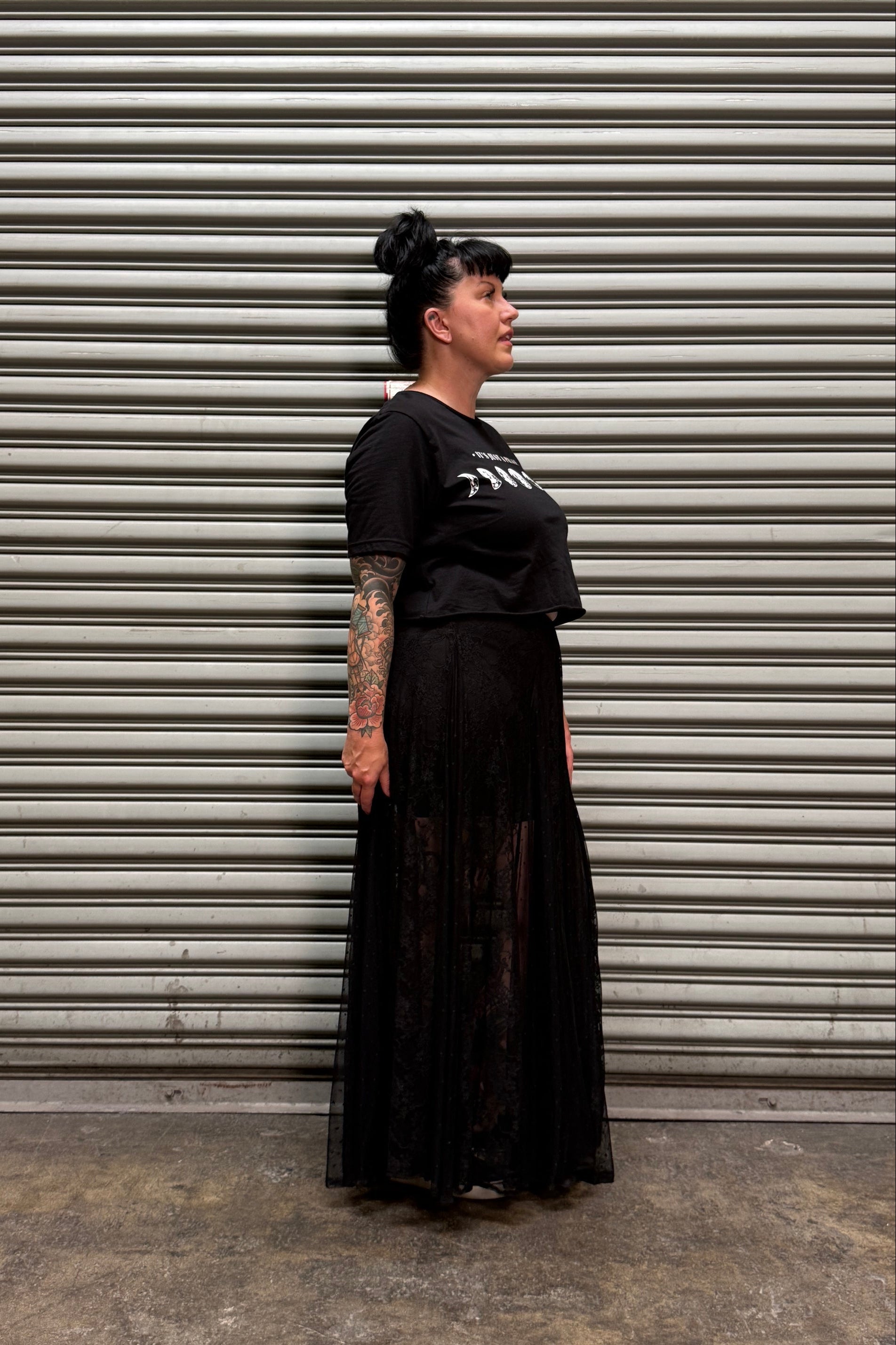 A woman with dark hair in a bun stands in profile against a gray metal roll-up door, wearing a black short-sleeve graphic T-shirt and a long black pleated skirt with sheer lace overlay.