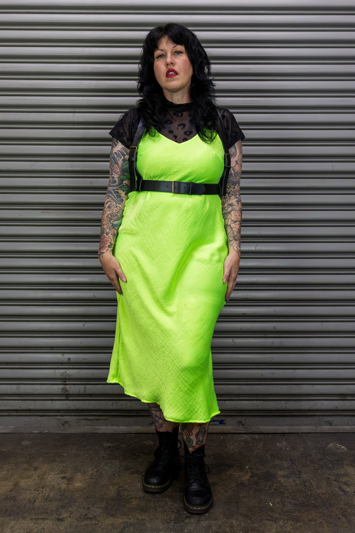 Person wearing a bright green dress with a black leather harness against a metal shutter background