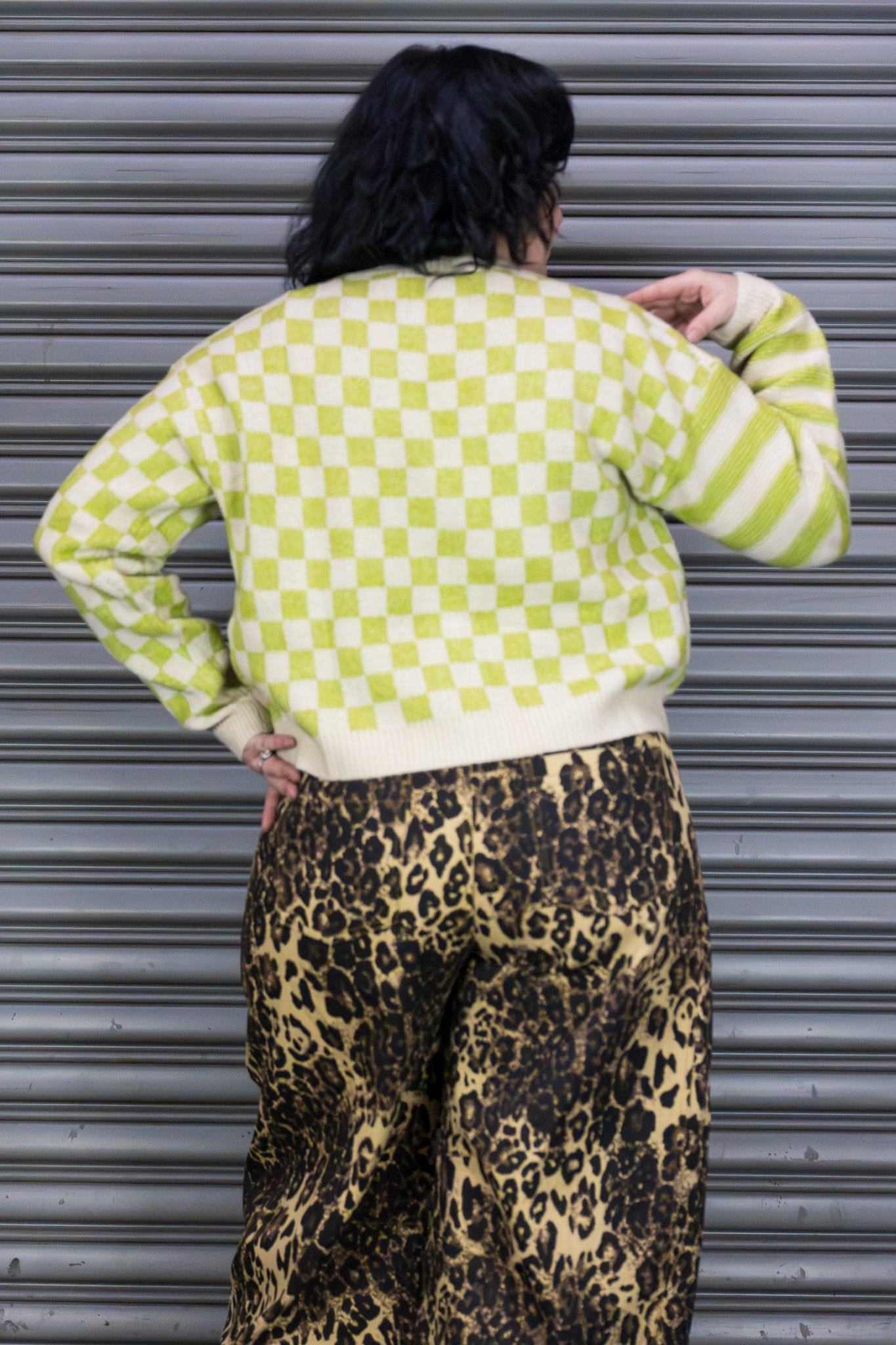 Person wearing a green and white checkered sweater and leopard print pants against a metallic shutter background.