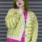 Person wearing a green and white checkered cardigan over a colorful shirt with a pink skirt, standing against a metallic shutter.