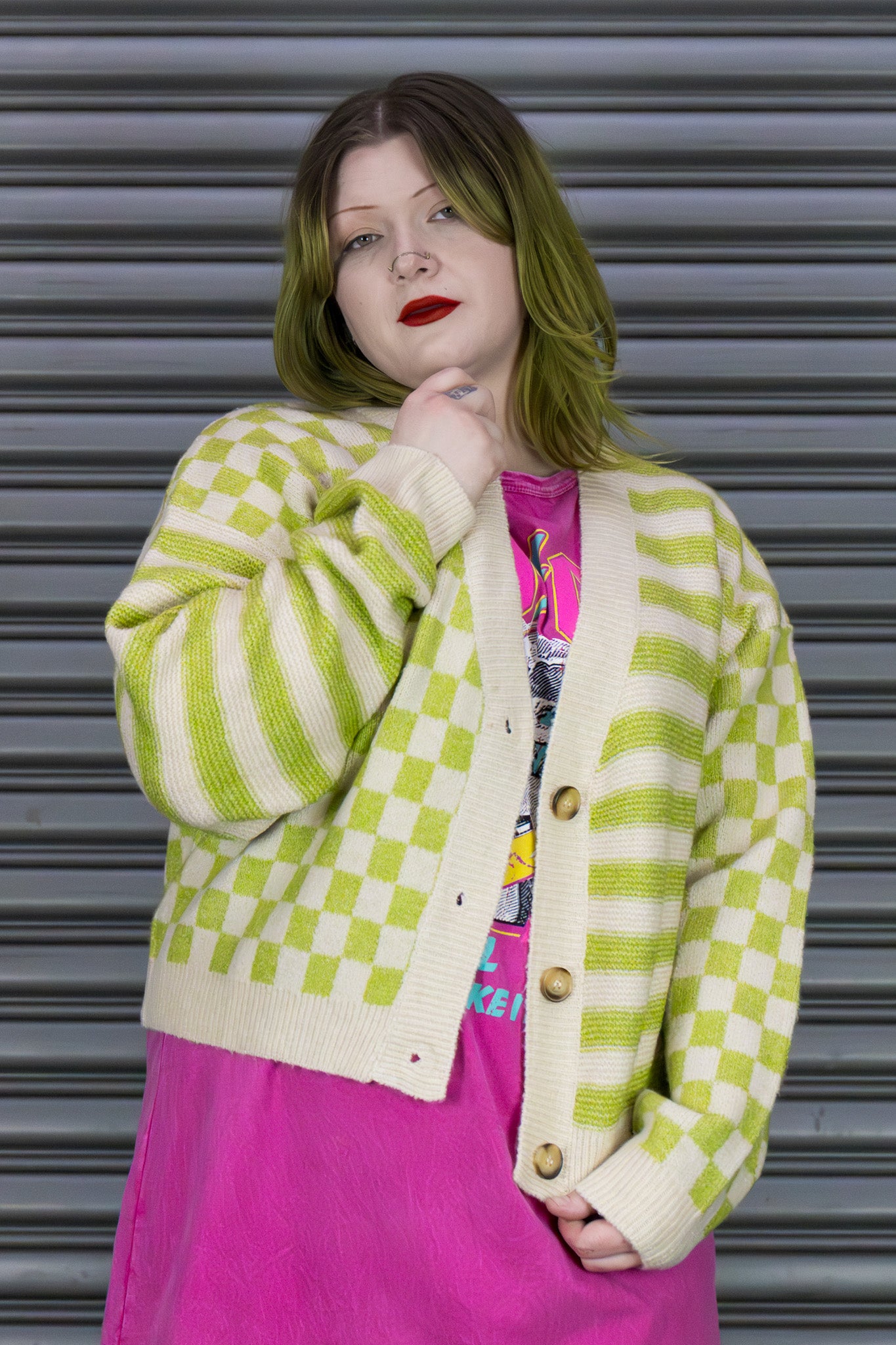 Person wearing a green and white checkered cardigan over a colorful shirt with a pink skirt, standing against a metallic shutter.