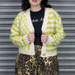 Person wearing a green and white checkered cardigan over a black shirt with leopard print pants, standing against a metallic shutter.