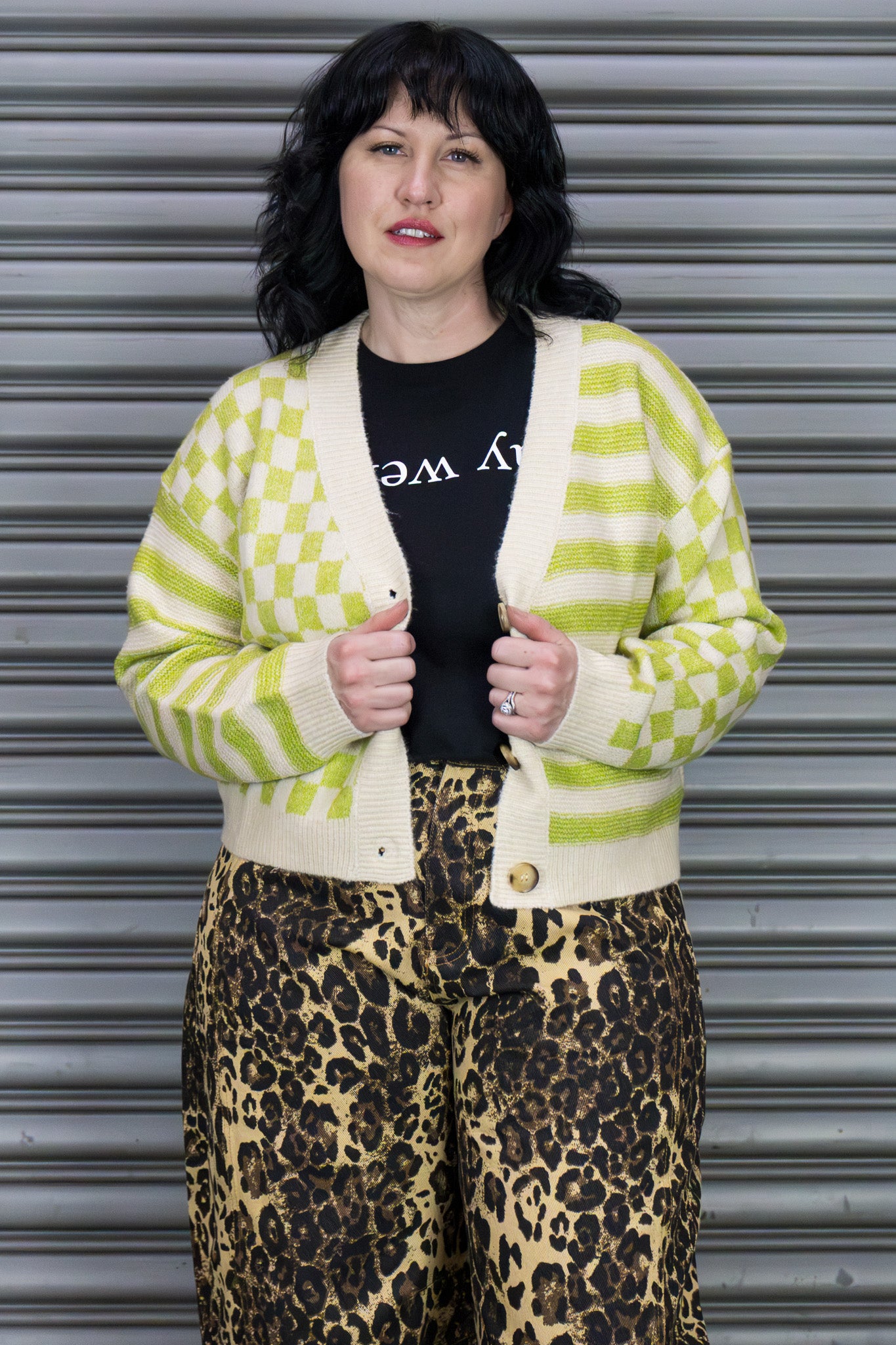 Person wearing a green and white checkered cardigan over a black shirt with leopard print pants, standing against a metallic shutter.