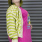 Person wearing a green and white checkered cardigan over a pink shirt with graphic designs, standing against a metallic shutter.