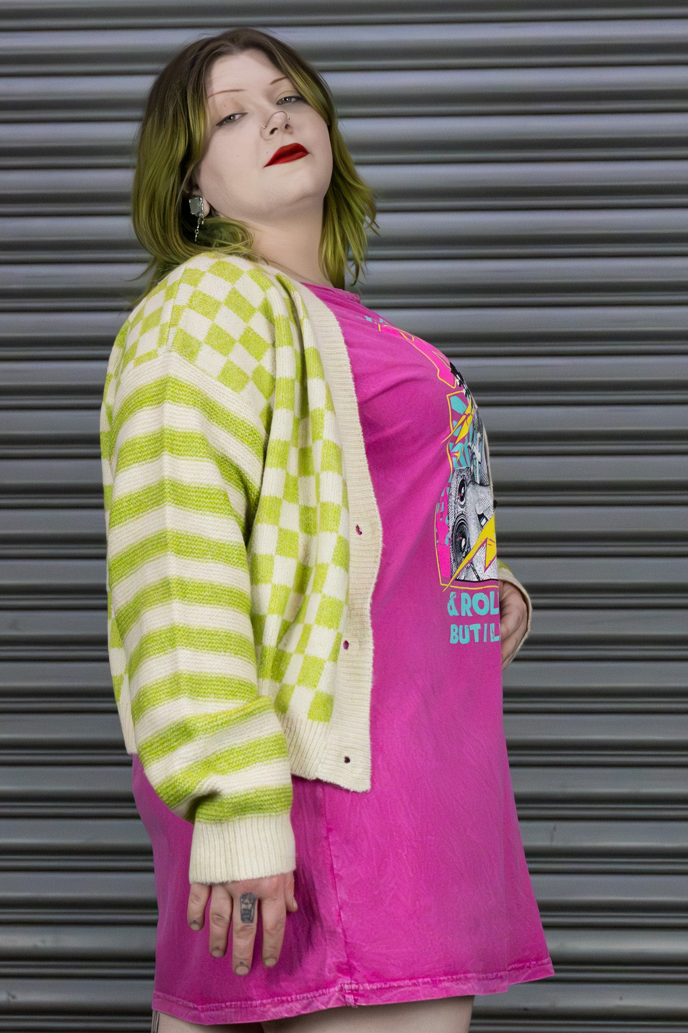 Person wearing a green and white checkered cardigan over a pink shirt with graphic designs, standing against a metallic shutter.