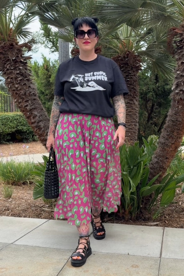 Person wearing a black t-shirt and pink floral skirt standing in front of palm trees.