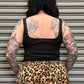 Person wearing a black tank top and leopard print skirt with tattoos on arms and back, standing against a metallic shutter.