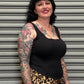 Woman with tattoos wearing a black top and leopard print pants standing in front of a gray shutter.