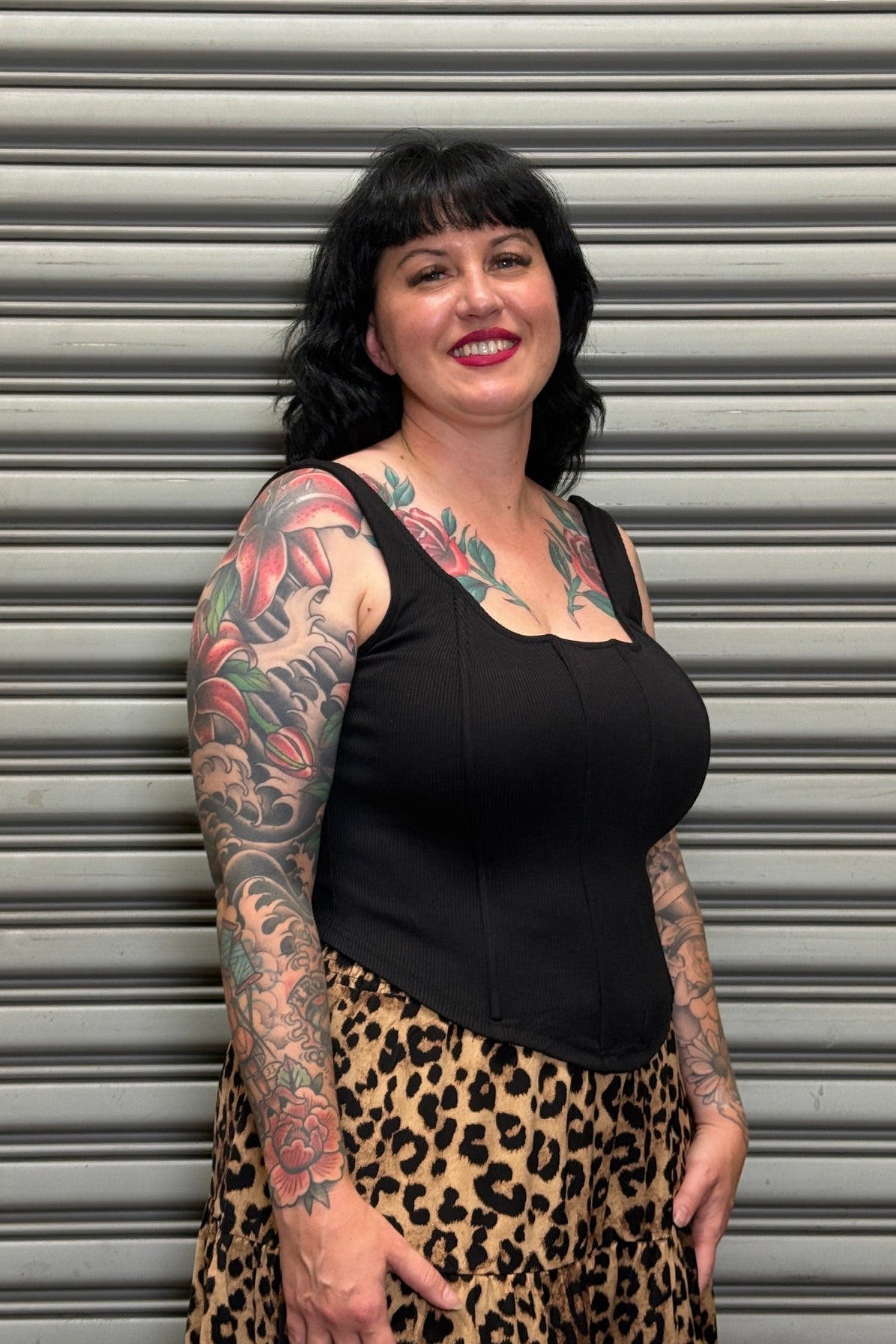 Woman with tattoos wearing a black top and leopard print pants standing in front of a gray shutter.