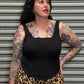 Woman wearing a black top and leopard print skirt with tattoos on arms and chest, standing against a metallic shutter.