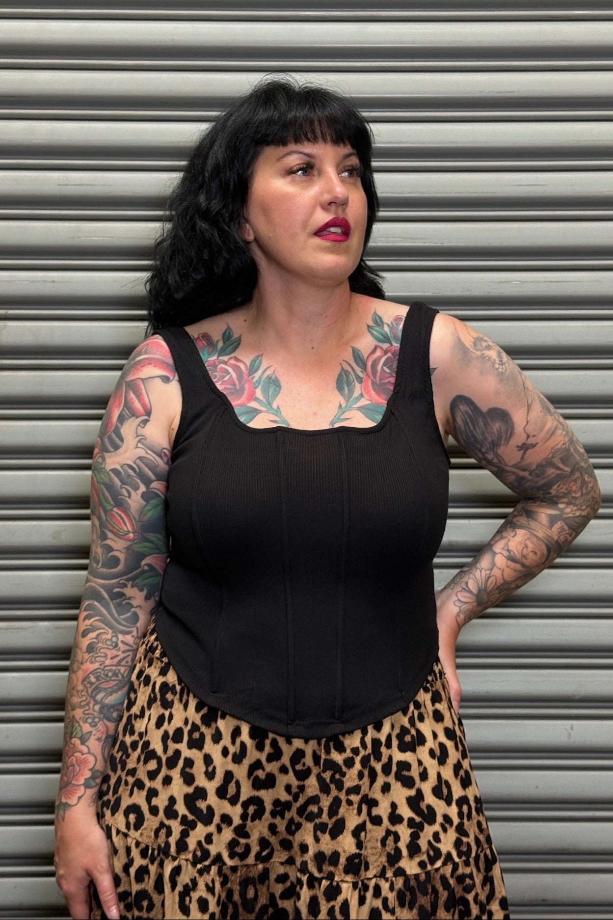 Woman wearing a black top and leopard print skirt with tattoos on arms and chest, standing against a metallic shutter.