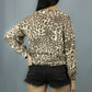 A woman shown from the back wearing a long-sleeve leopard print top and black denim shorts against a plain gray studio background.