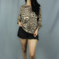 A woman modeling a beige and brown leopard print long-sleeve top with black denim shorts, standing against a gray studio backdrop.