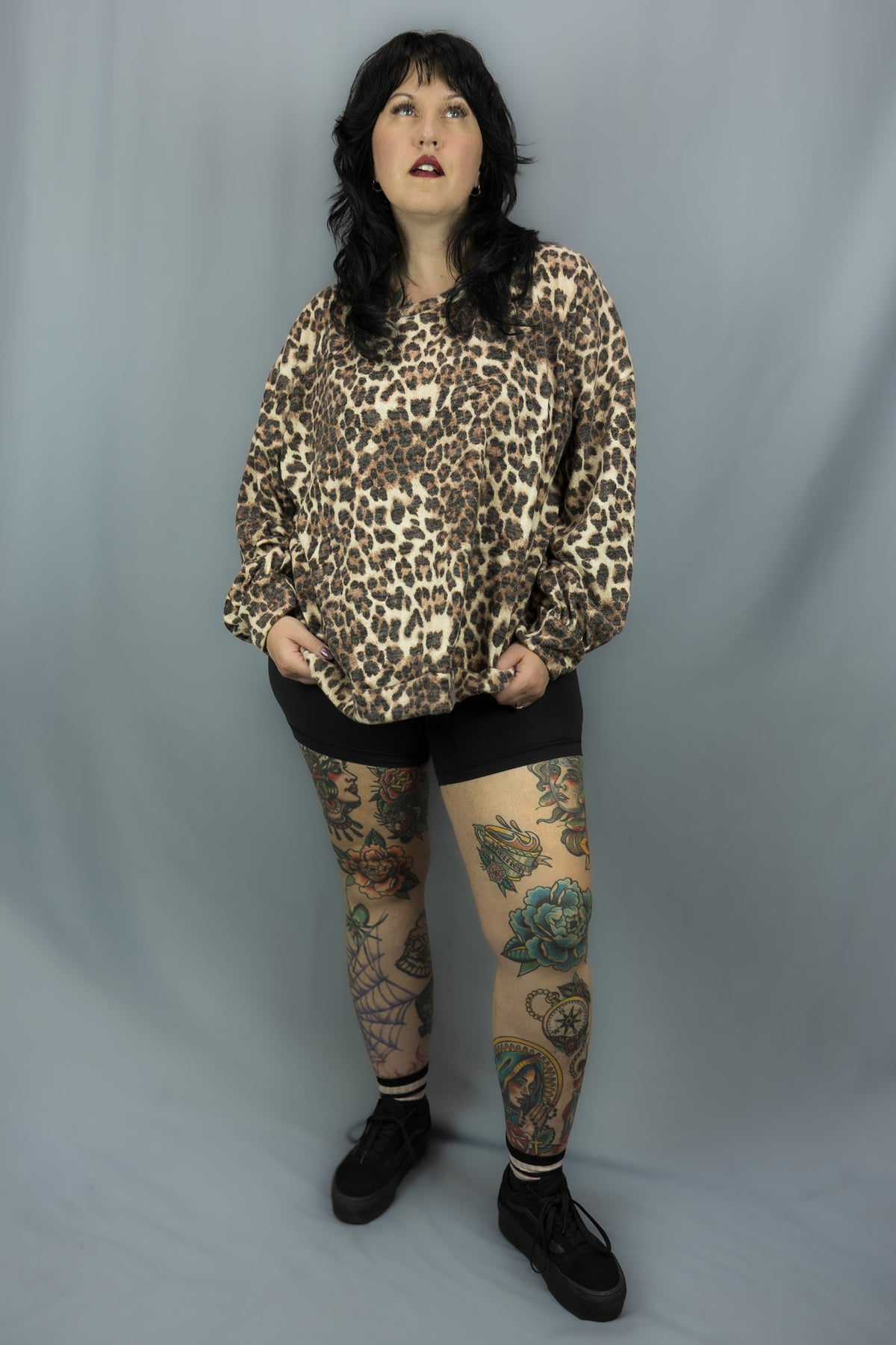 A woman modeling a leopard print long-sleeve top, black shorts, and black lace-up shoes against a gray studio backdrop.