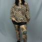 A woman modeling a leopard print long-sleeve sweatshirt, black shorts, and black tights with colorful leg tattoos, standing against a gray studio backdrop.