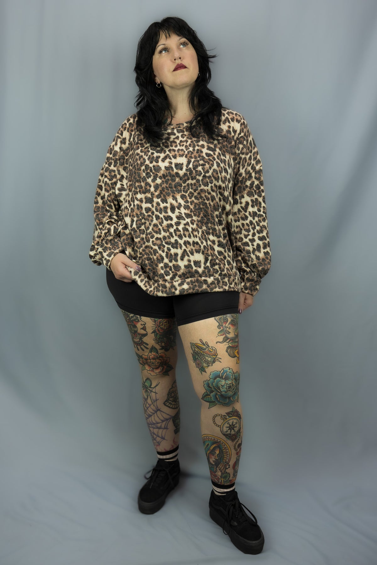 A woman modeling a leopard print long-sleeve sweatshirt, black shorts, and black tights with colorful leg tattoos, standing against a gray studio backdrop.