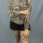 A woman wearing a beige and brown leopard print long-sleeve top with black frayed denim shorts, standing against a solid gray studio backdrop.
