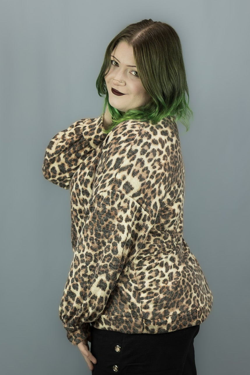 A person wearing a beige and brown leopard print jacket and black wide-leg pants with side grommets, standing against a gray studio backdrop.