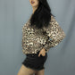 A woman posing in a leopard print long-sleeve top and black denim shorts against a solid gray studio background.