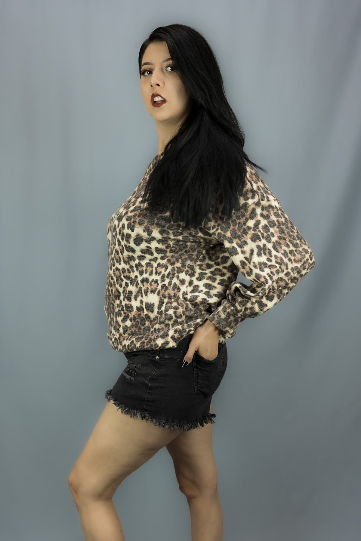 A woman posing in a leopard print long-sleeve top and black denim shorts against a solid gray studio background.